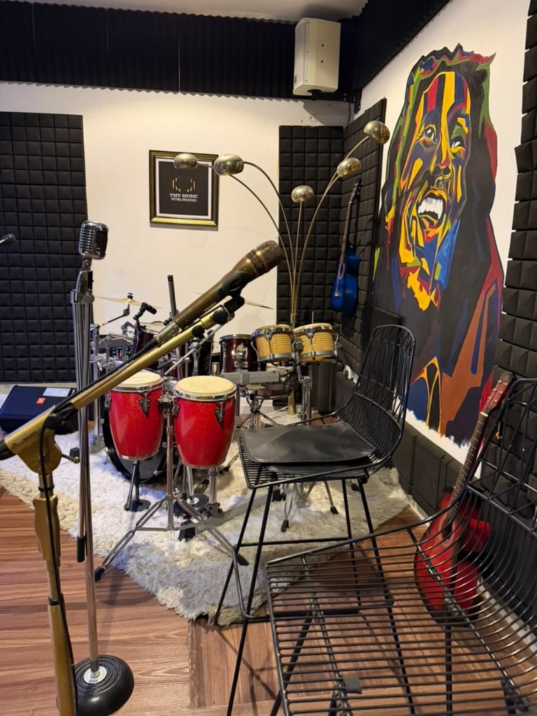 Rehearsal corner with congas, a wire chair, guitars, and the mural.