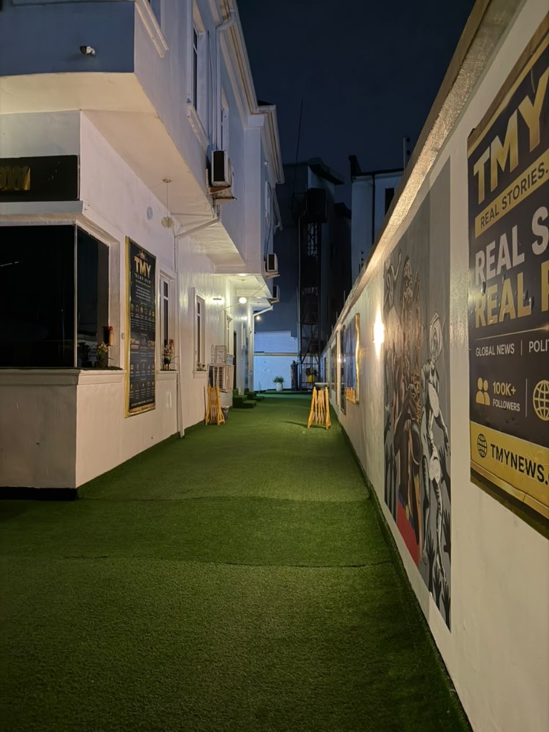 Night walkway beside the building, with green turf and the TMY News wall.